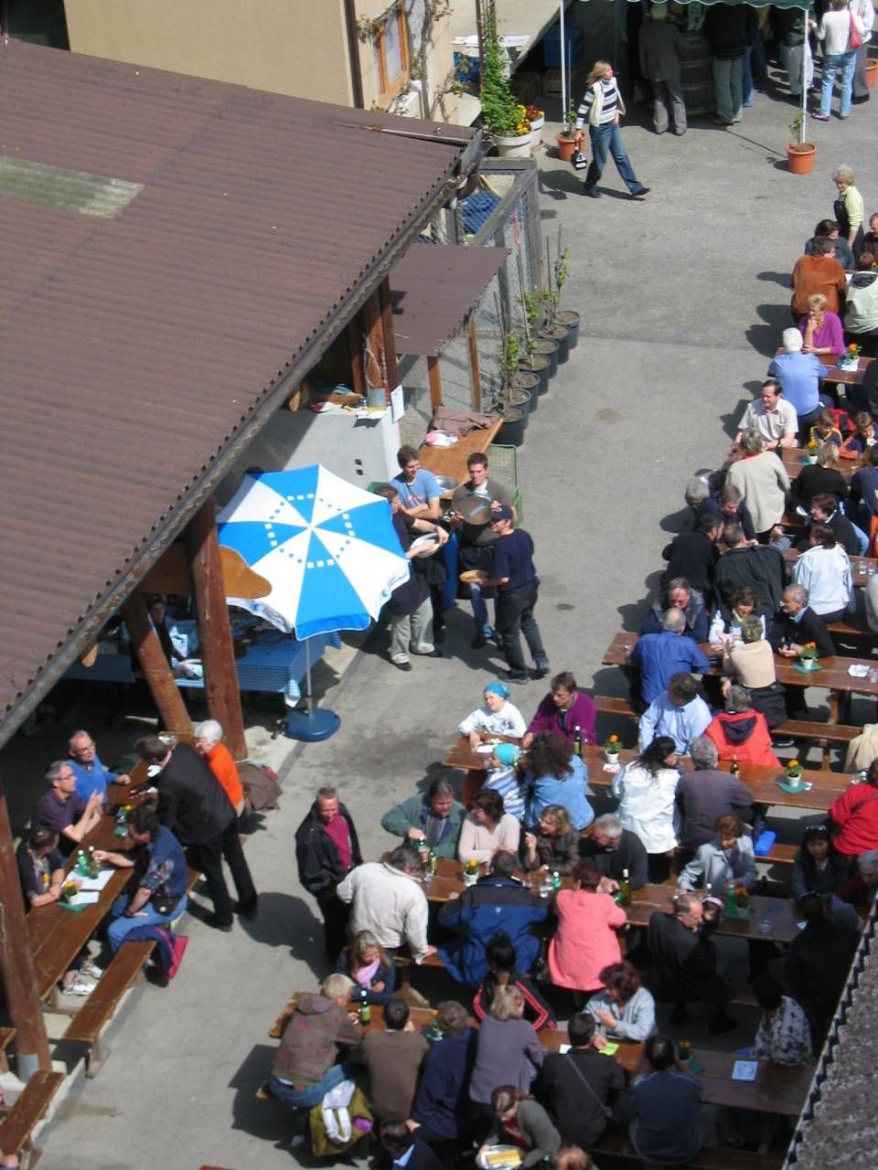 Eine Menschengruppe sitzt im Freien an Tischen unter einer braunen Markise und einem blau-weissen Sonnenschirm.