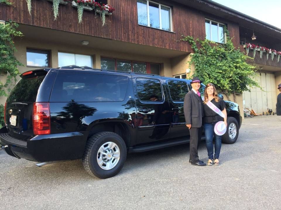 Zwei Personen stehen neben einem schwarzen Geländewagen. Sie sind festlich gekleidet, vor einem Gebäude mit Grünanlagen.