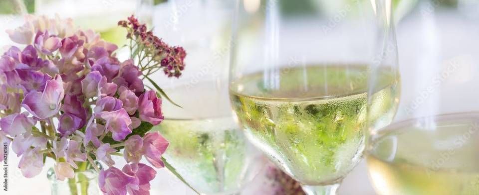 Weingläser mit gelber Flüssigkeit vor einem Strauss rosa und violetter Blumen.