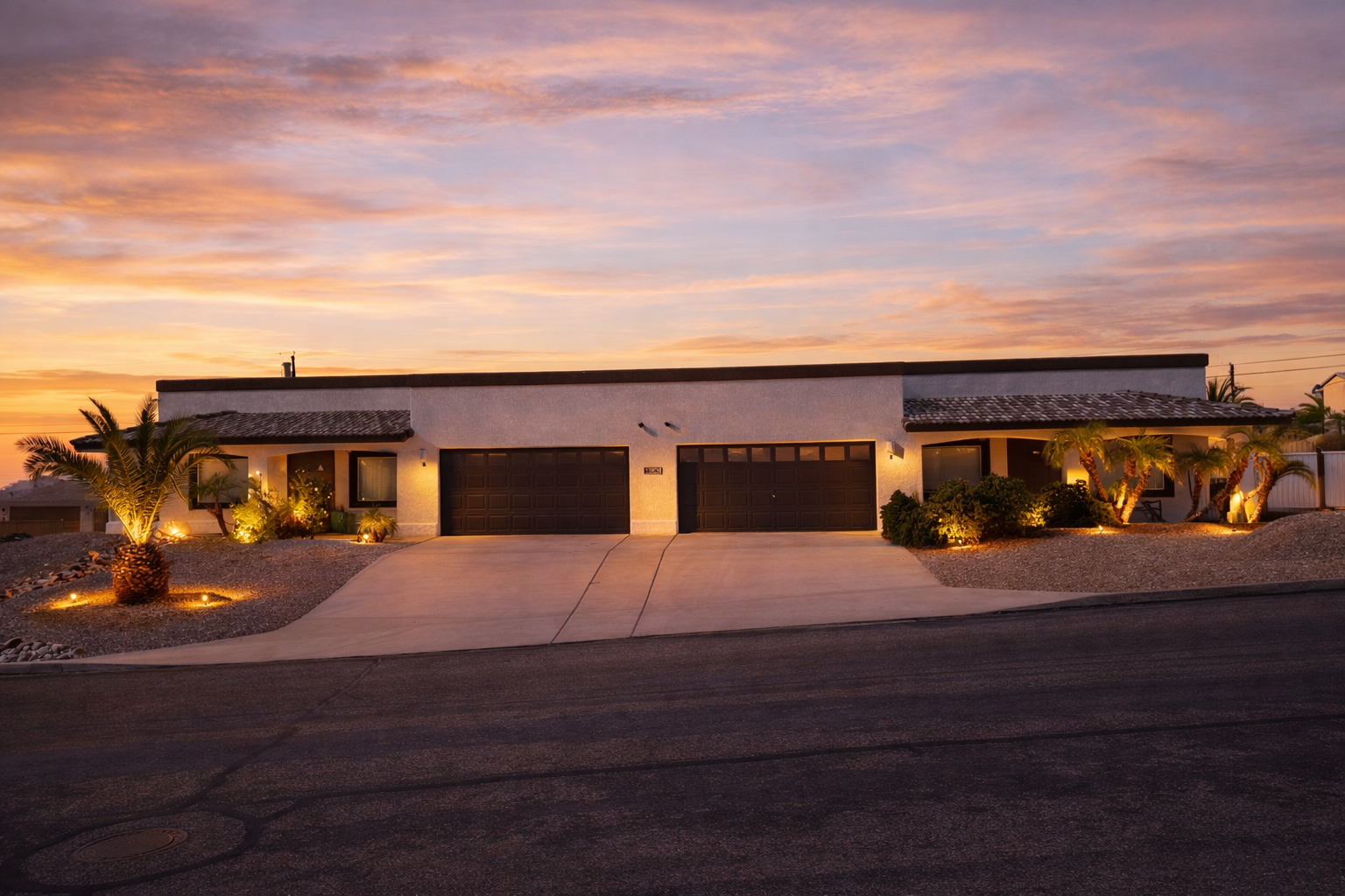 White house with two dark garage doors, palm trees lit by landscape lighting, and sunset sky.