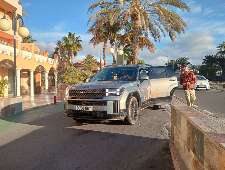 Un SUV plateado en una calle soleada de un complejo turístico, con palmeras y una persona de pie junto a la acera.