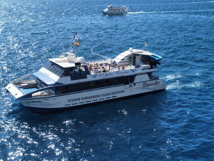 Un ferry blanco de pasajeros con gente en cubierta navegando por el océano azul; un barco más pequeño se divisa a lo lejos.