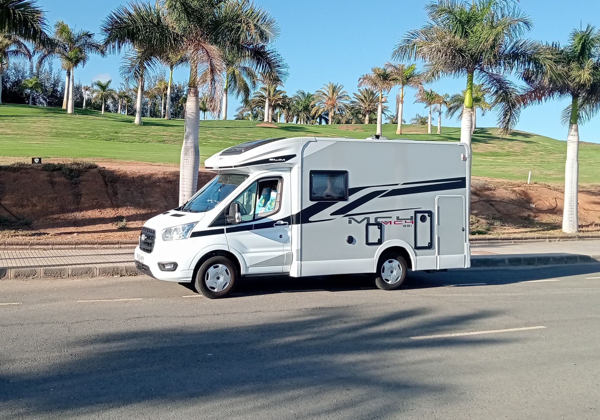 Una autocaravana blanca aparcada en una carretera junto a palmeras y un terraplén cubierto de hierba.