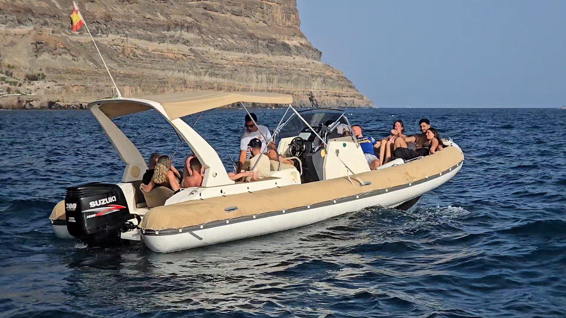 Gente paseando en una lancha neumática de color beige sobre aguas azules cerca de una costa rocosa.