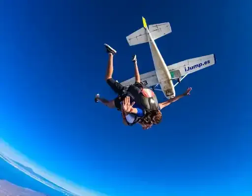 Dos paracaidistas en tándem caen en caída libre junto a un avión azul y blanco bajo un cielo azul despejado.