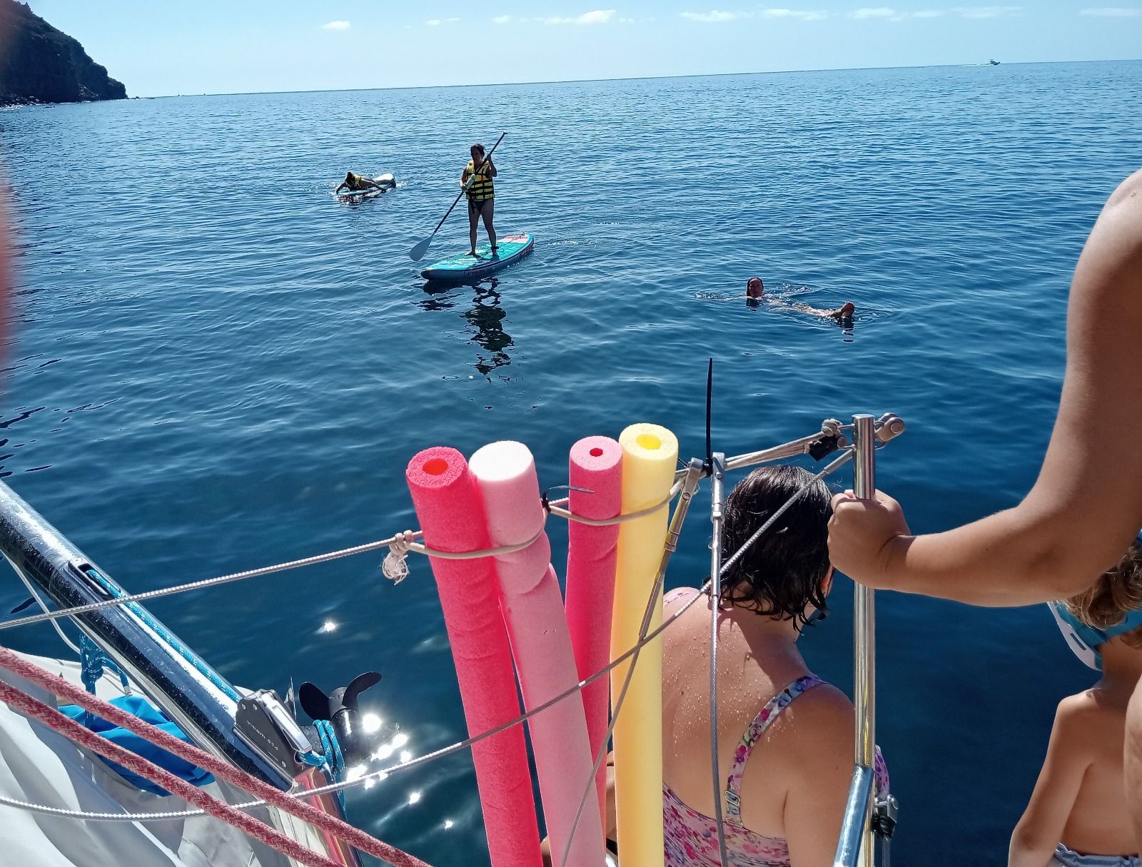 Gente en un barco observando a practicantes de paddleboard en las tranquilas aguas azules del océano, cerca de coloridos flotadores de espuma para piscinas.