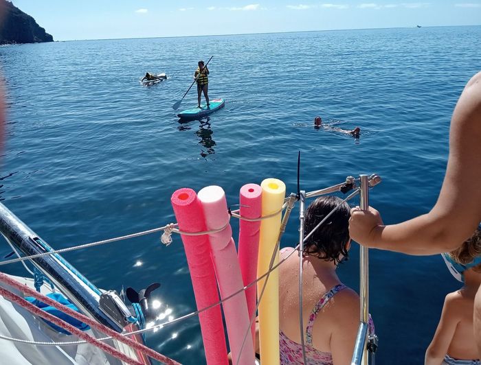 Gente en un barco observando a practicantes de paddleboard en las tranquilas aguas azules del océano, cerca de coloridos flotadores de espuma para piscinas.