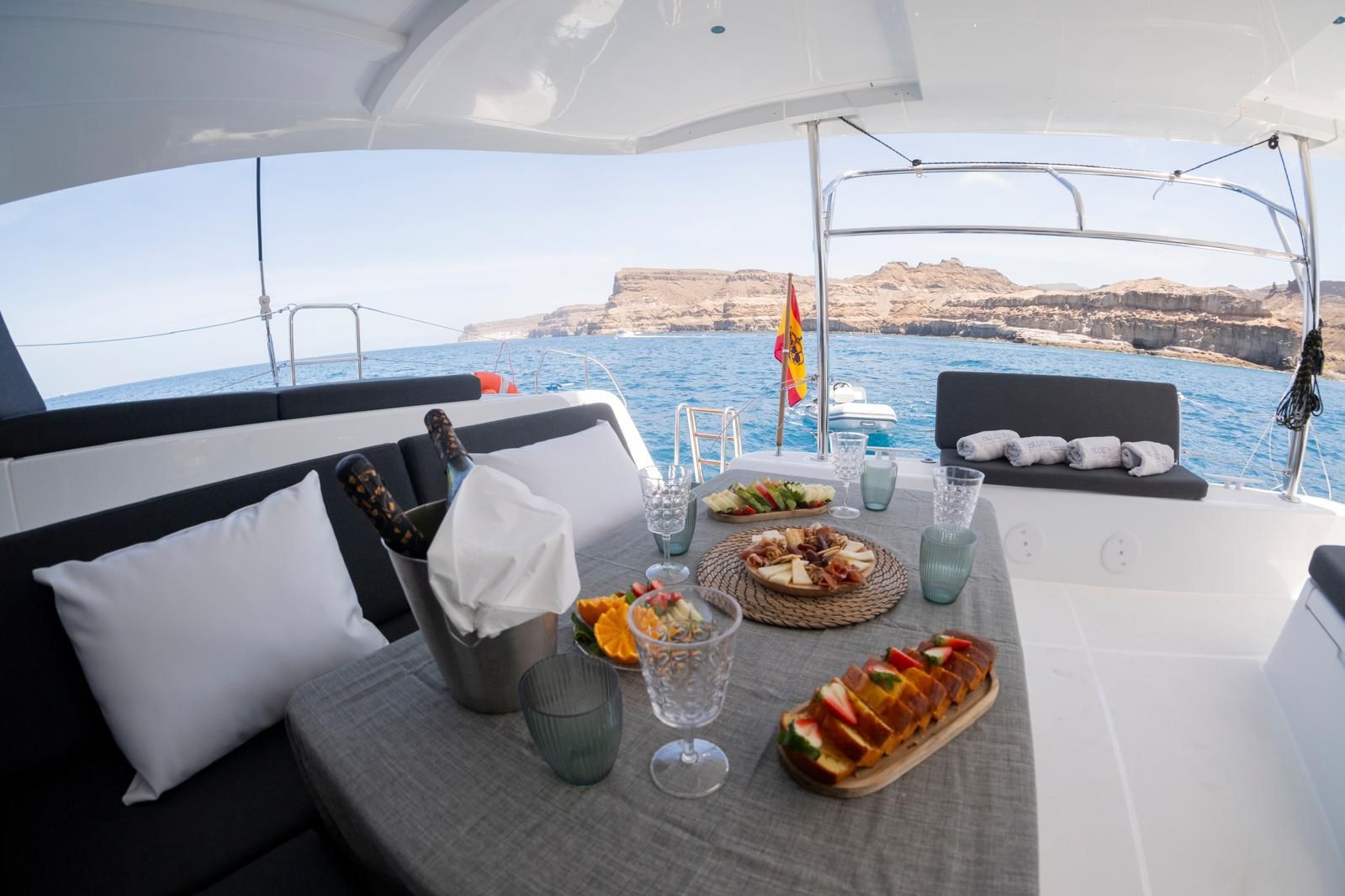 Mesa de comedor en un yate con vistas soleadas al mar, con comida, bebidas y cojines bajo un toldo.