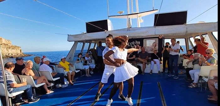Bailarines actuando en la cubierta de un barco con pasajeros sentados observando bajo un cielo azul brillante.