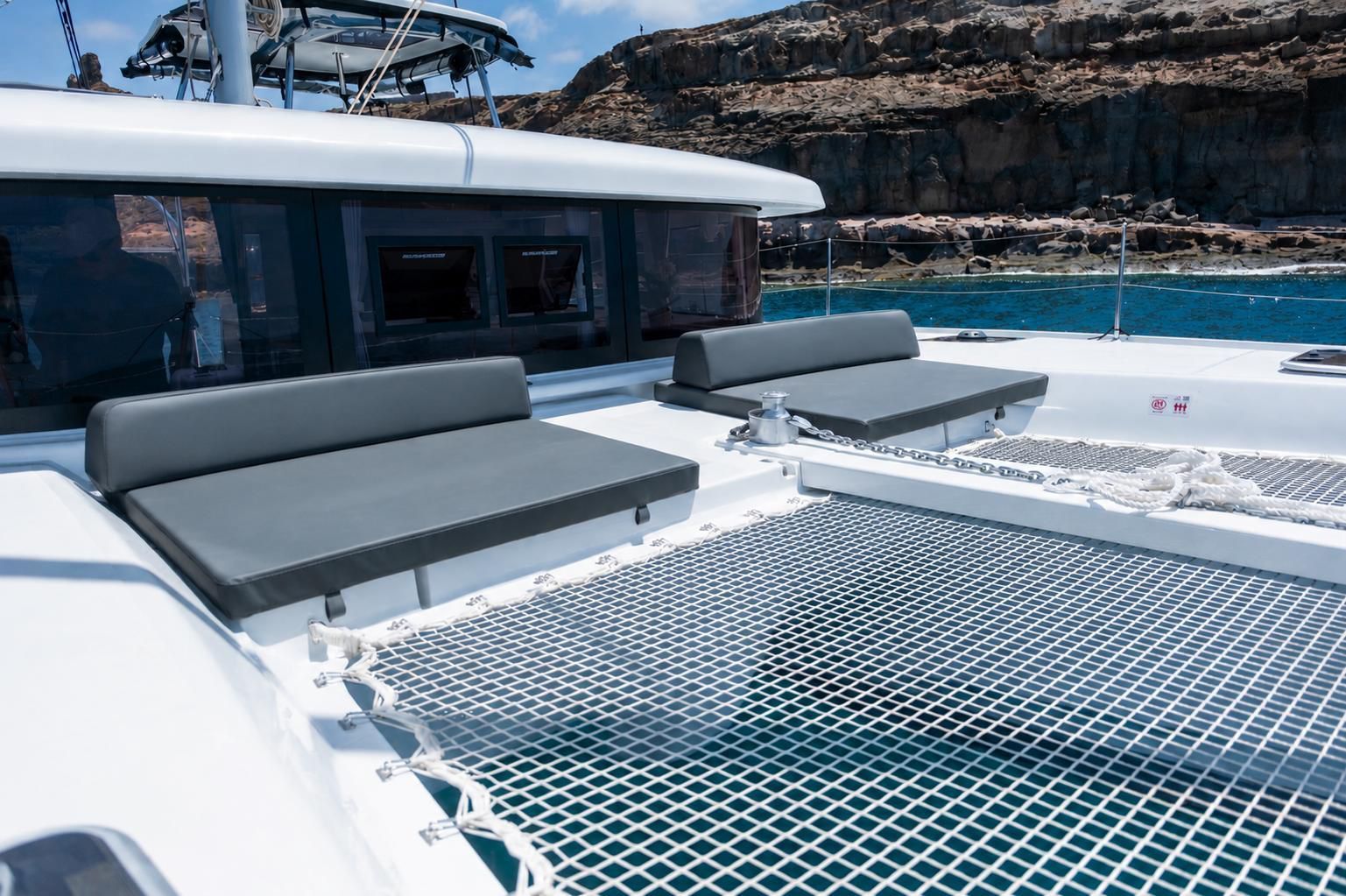 Cubierta de catamarán de lujo con tumbonas grises y un trampolín con red en aguas azules tranquilas cerca de la costa rocosa.