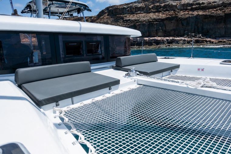 Cubierta de catamarán de lujo con tumbonas grises y un trampolín con red en aguas azules tranquilas cerca de la costa rocosa.