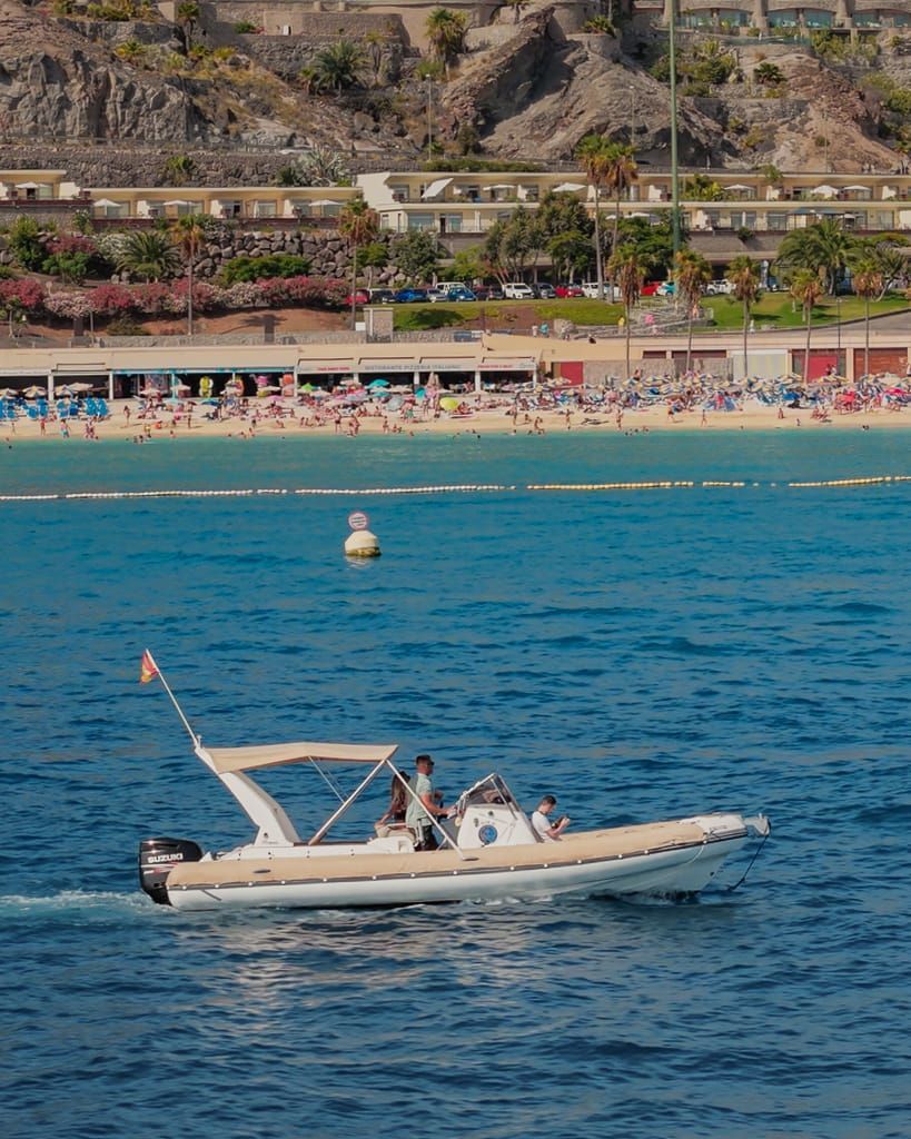 Lancha motora blanca con dos personas a bordo en aguas azules cerca de una playa concurrida y una ladera rocosa.