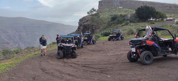 Vehículos todoterreno estacionados en un mirador de tierra junto a un cañón, con gente reunida bajo un cielo nublado.
