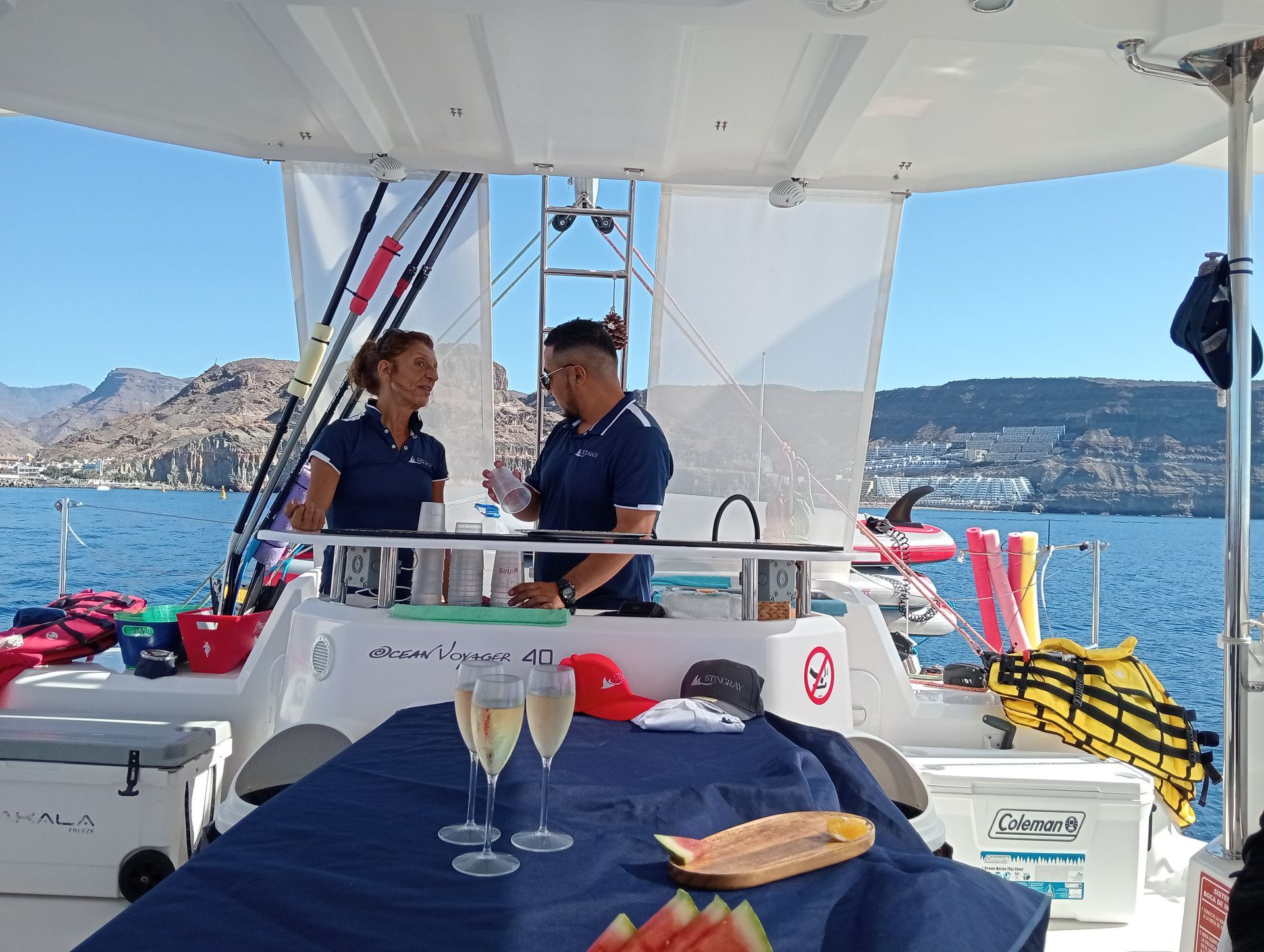 Dos personas en la cubierta de un barco, tomando algo, bajo un toldo blanco, con vistas a una bahía azul y a las colinas lejanas.