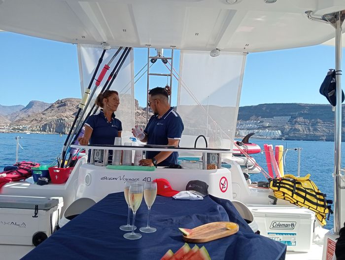 Dos personas en la cubierta de un barco, tomando algo, bajo un toldo blanco, con vistas a una bahía azul y a las colinas lejanas.