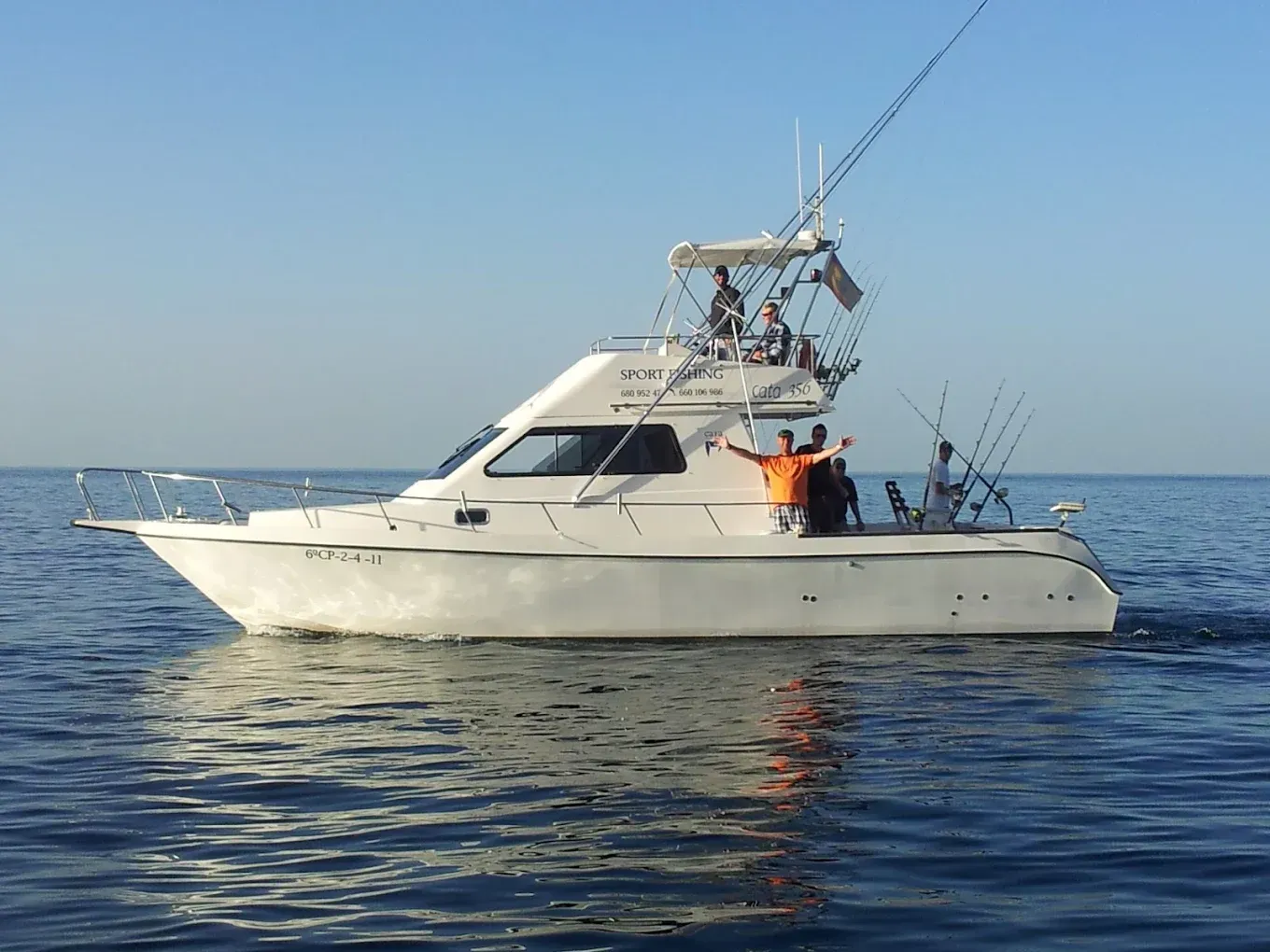 Un barco de pesca blanco navega en aguas azules y tranquilas, con dos personas en cubierta y las cañas de pescar levantadas.