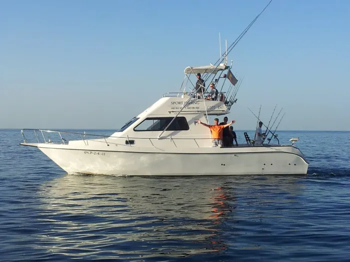 Un barco de pesca blanco navega en aguas azules y tranquilas, con dos personas en cubierta y las cañas de pescar levantadas.