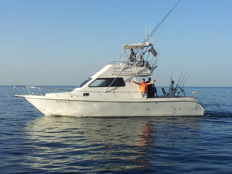 Un barco de pesca blanco navega en aguas azules y tranquilas, con dos personas en cubierta y las cañas de pescar levantadas.