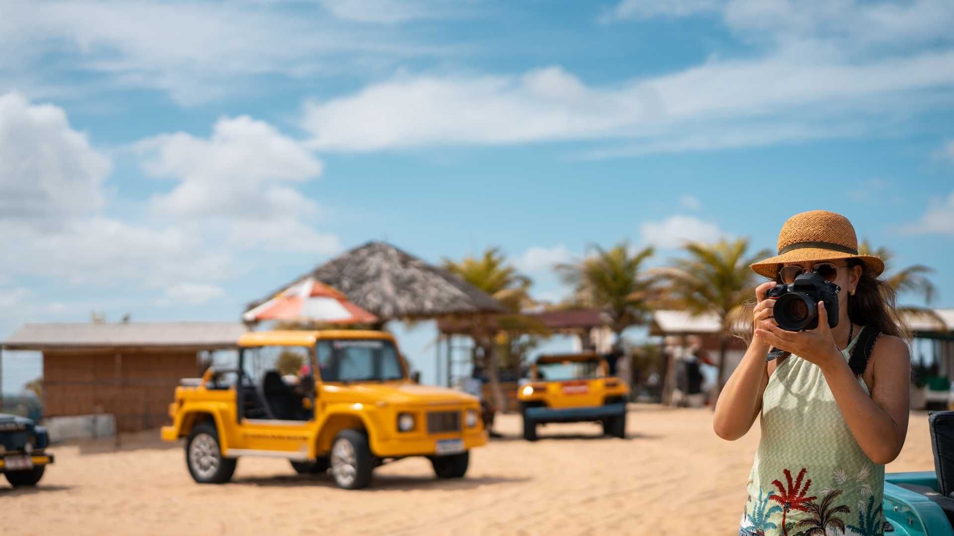 Mujer tomando una foto cerca de jeeps amarillos en una playa con palmeras y un cielo azul.