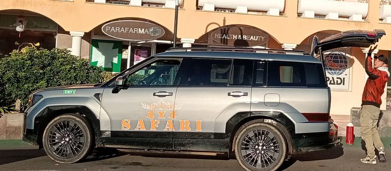 Un SUV plateado con letras naranjas estacionado frente a una tienda, con una persona de pie a su lado.