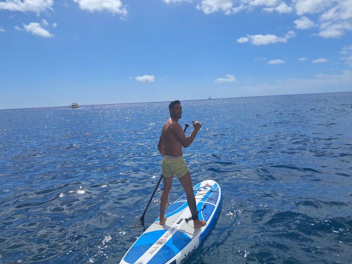 Hombre de pie sobre una tabla de remo azul en mar abierto bajo un cielo soleado.