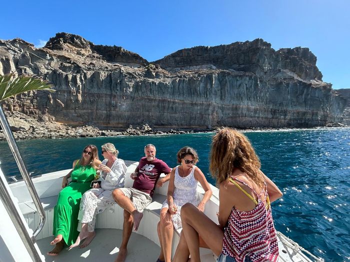 Gente relajándose en un barco en aguas azules con acantilados rocosos al fondo.