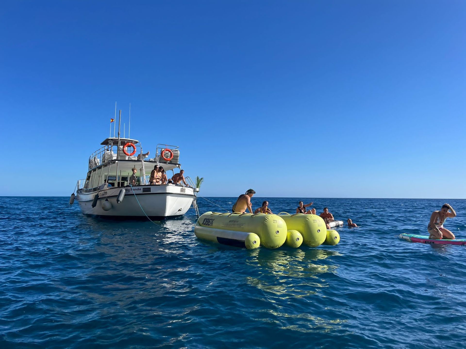Barco y gente en el océano azul, con una colchoneta inflable amarilla flotando cerca bajo un cielo despejado.