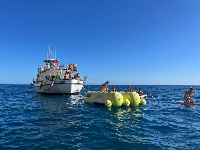 Barco y gente en el océano azul, con una colchoneta inflable amarilla flotando cerca bajo un cielo despejado.