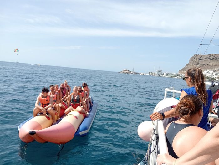 Personas paseando en una banana inflable roja en el mar, remolcada cerca de una costa rocosa.