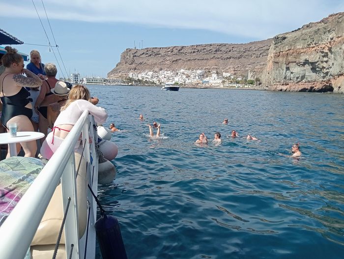 Gente nadando junto a un barco cerca de una costa rocosa bajo un cielo azul despejado.