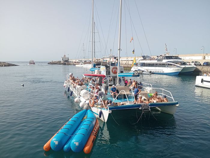Gente a bordo de un catamarán con balsas inflables en un puerto deportivo tranquilo, rodeados de otros barcos y yates.