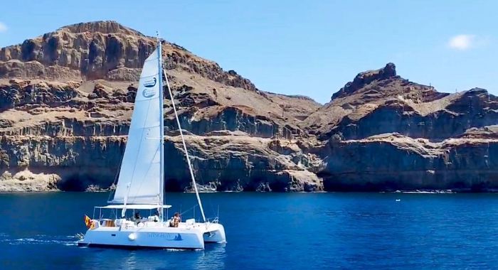 Velero blanco sobre aguas azules frente a acantilados rocosos costeros bajo un cielo despejado.