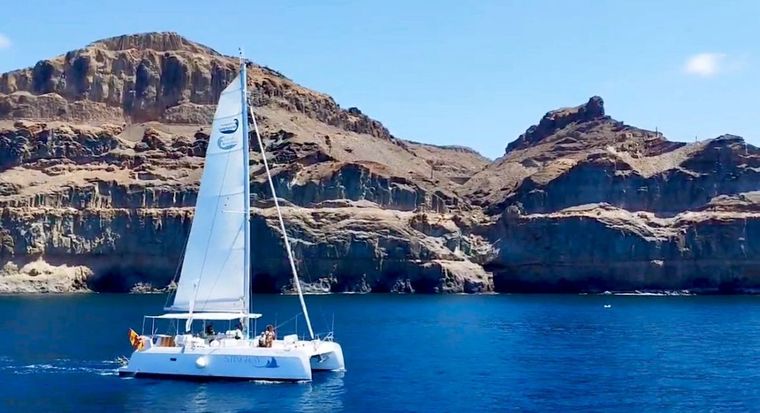 Velero blanco sobre aguas azules frente a acantilados rocosos costeros bajo un cielo despejado.