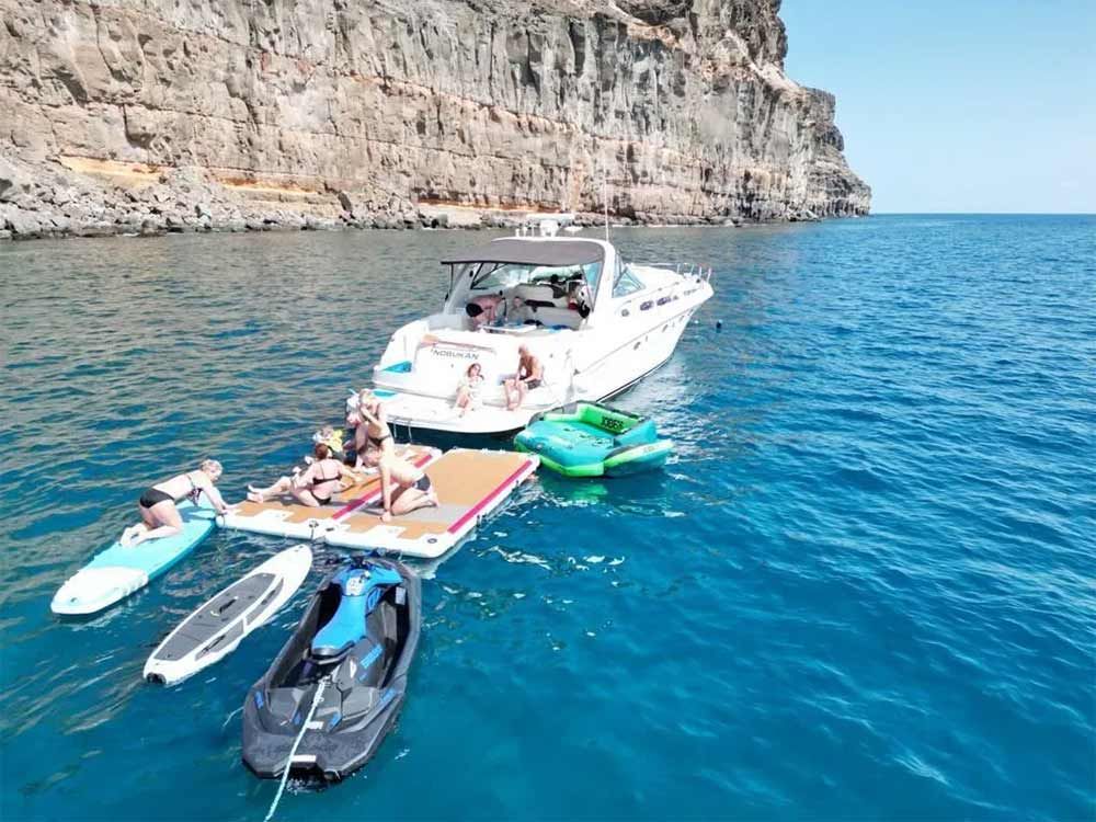 Gente relajándose en un yate con kayaks y tablas de paddle surf junto a un acantilado rocoso costero.