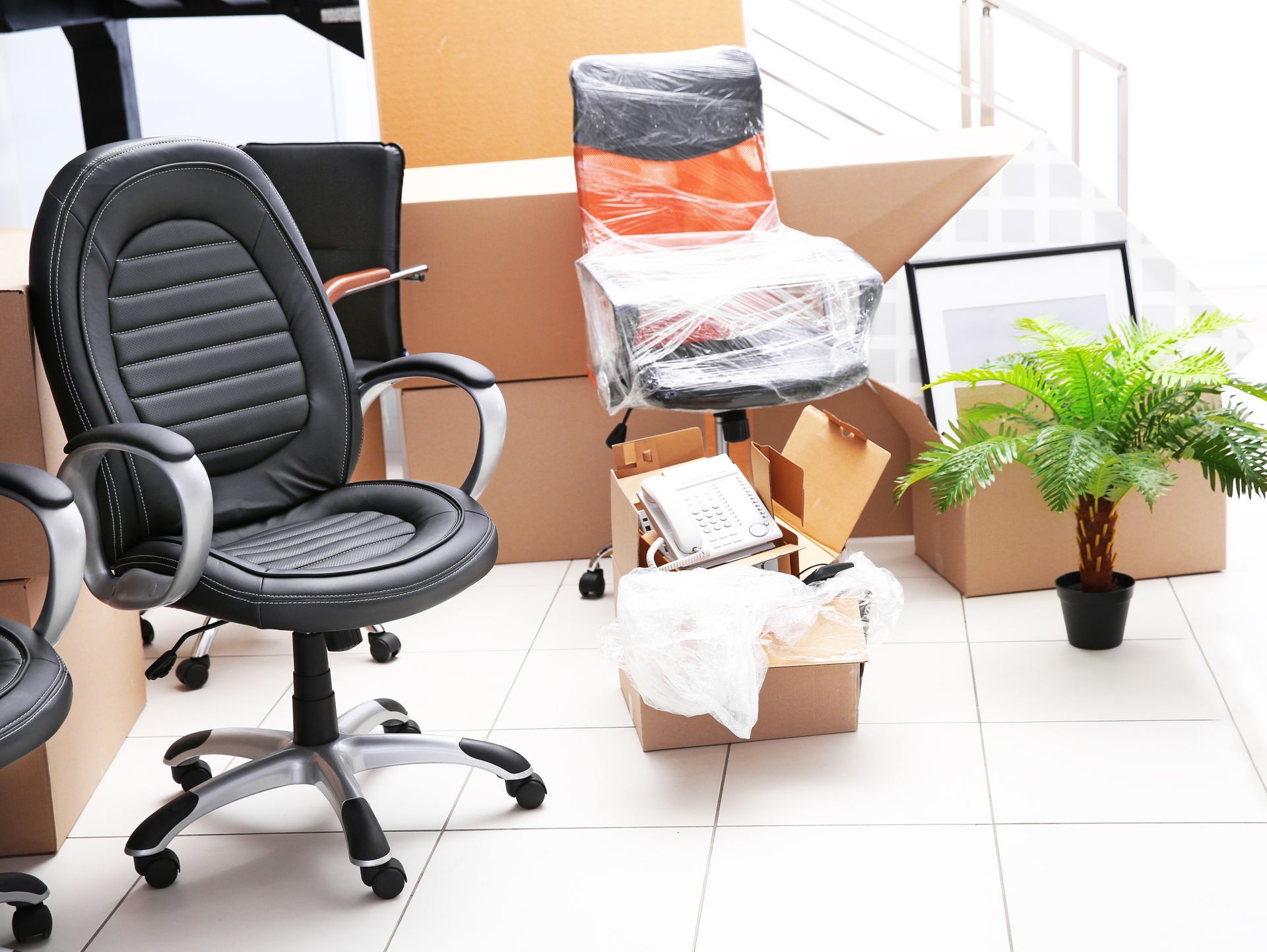 Bureau avec une chaise pivotante noire, des chaises emballées empilées dans des cartons et une plante en pot sur un sol carrelé.