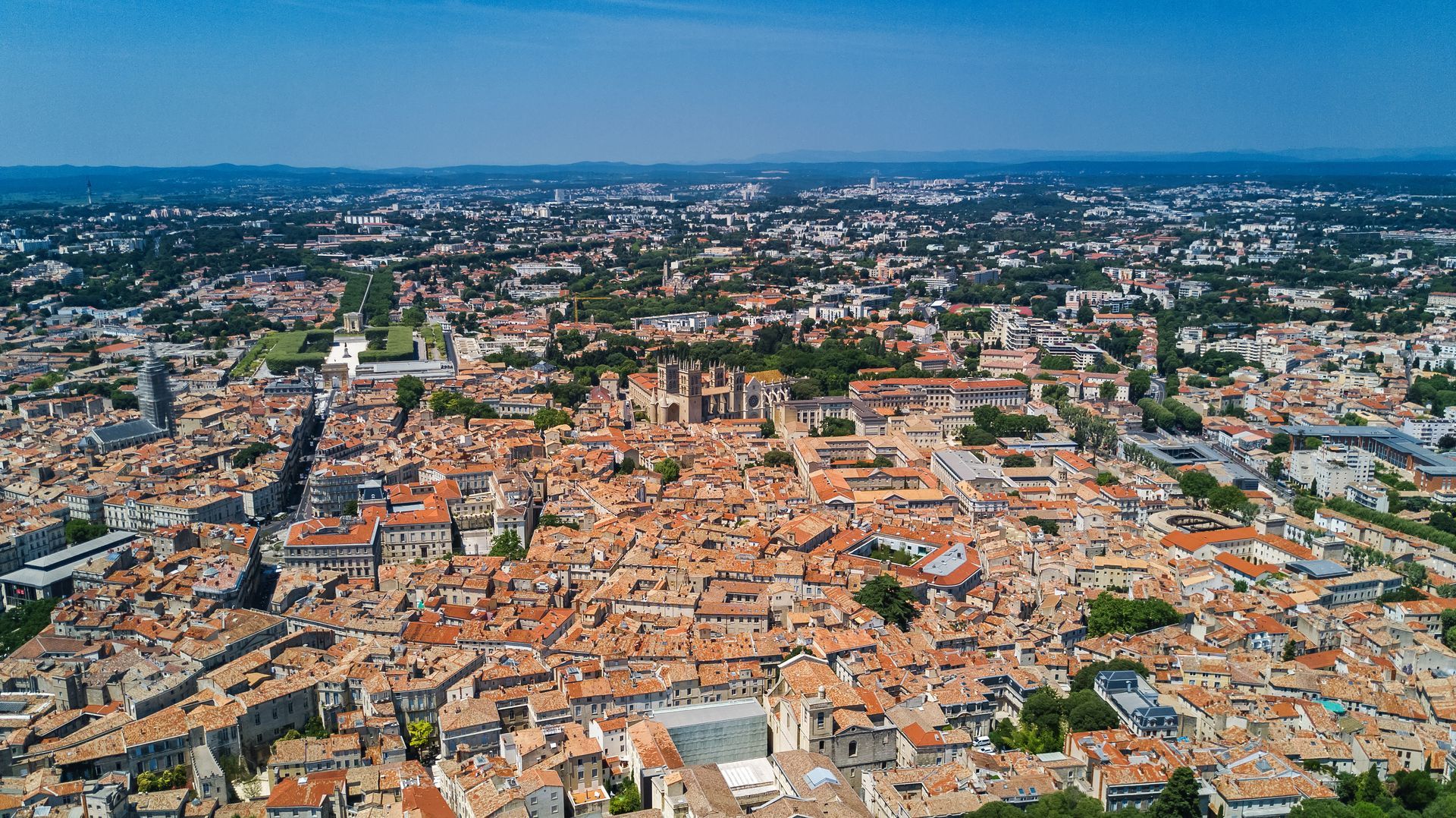 Vue aérienne d'une ville dense aux toits orangés s'étendant jusqu'à l'horizon sous un ciel bleu.
