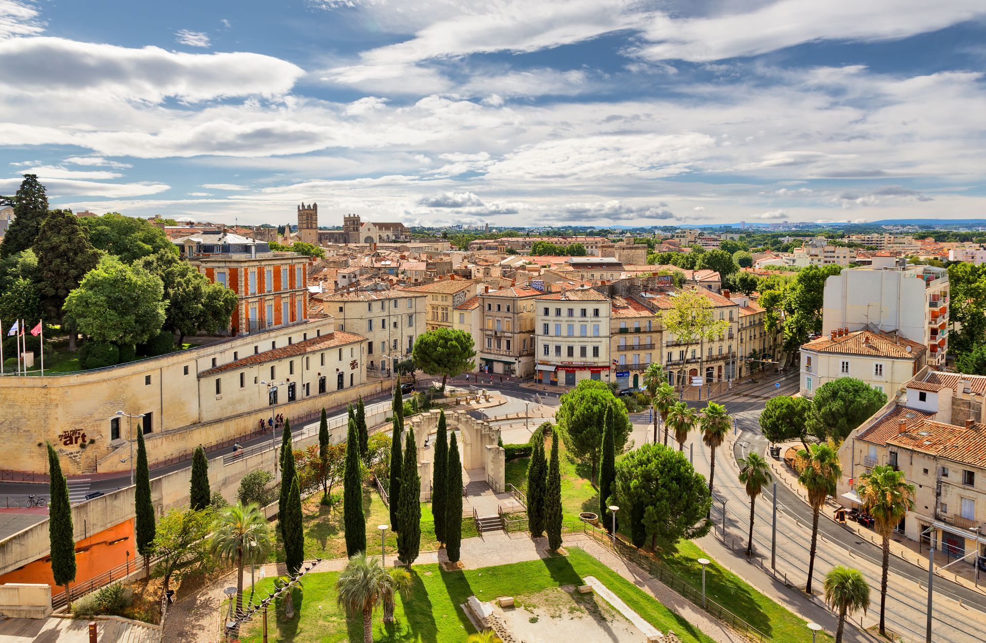 Vue panoramique d'une ville historique aux toits de tuiles rouges, aux arbres et sous un ciel nuageux, depuis un belvédère à flanc de colline.