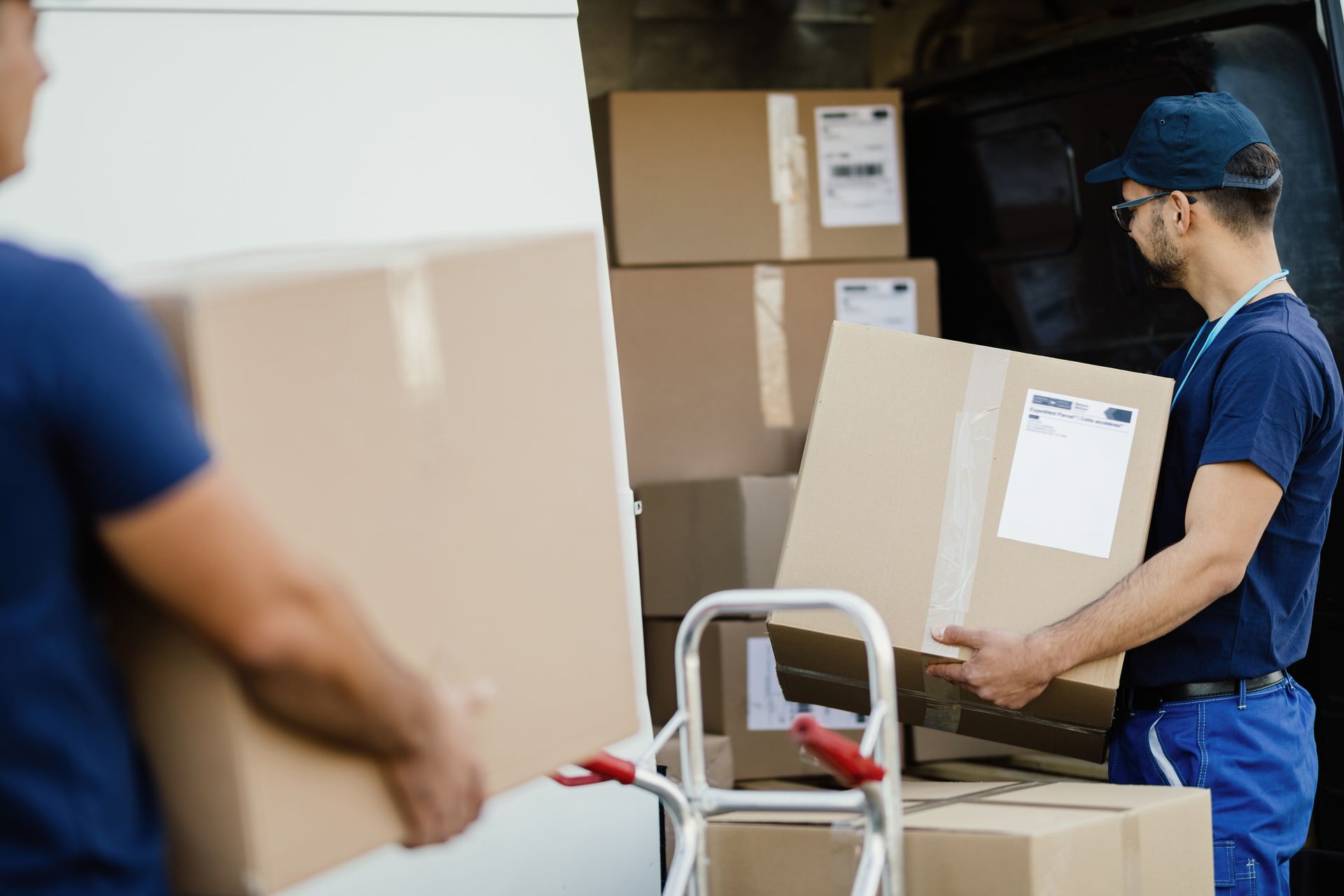 Un employé d'entrepôt en uniforme bleu déplace des cartons à l'aide d'un diable dans une zone de stockage.