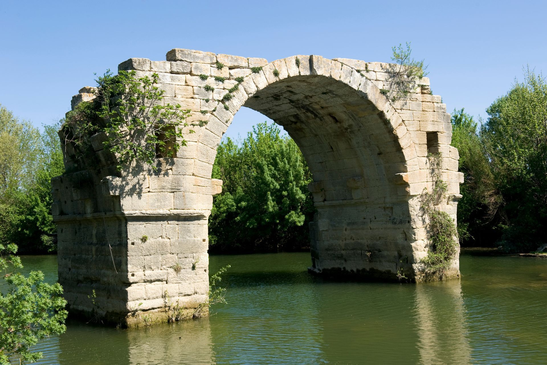 Ruines d'un pont en arc de pierre enjambant une rivière verdoyante et paisible, entourées d'arbres sous un ciel bleu.