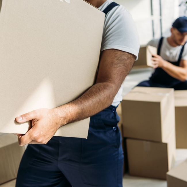 Deux déménageurs transportent de grands cartons dans un entrepôt ou une zone de déménagement.