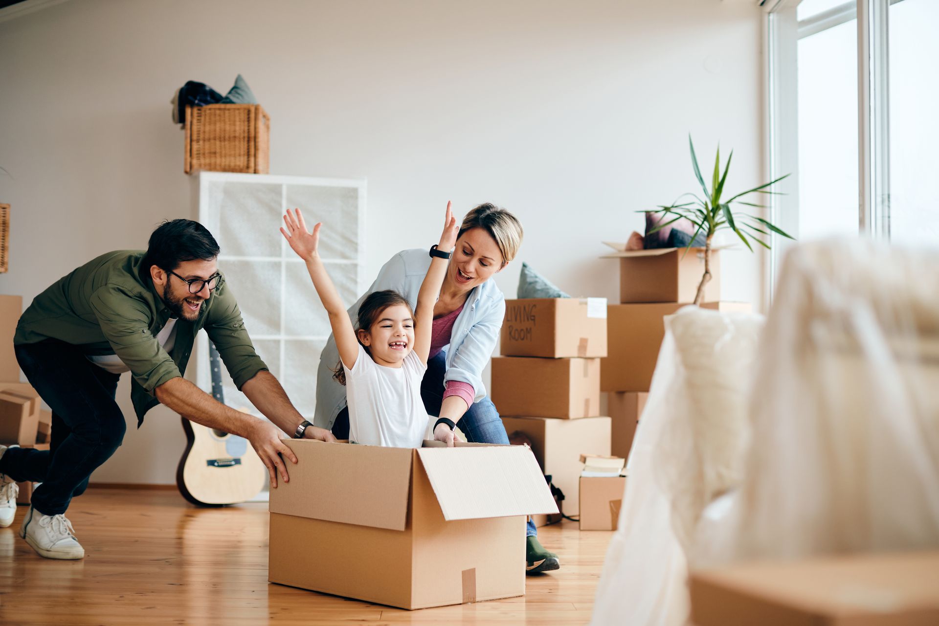 Une famille fait ses cartons dans un salon lumineux lors d'un déménagement, un enfant applaudissant dans un carton ouvert.