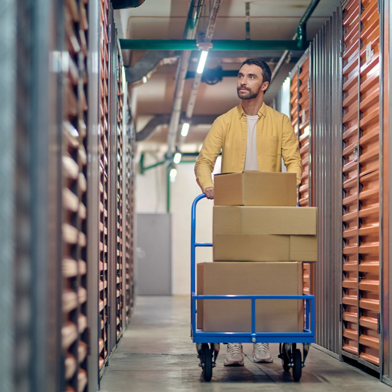 Un homme pousse un chariot bleu rempli de cartons entre les allées de stockage.