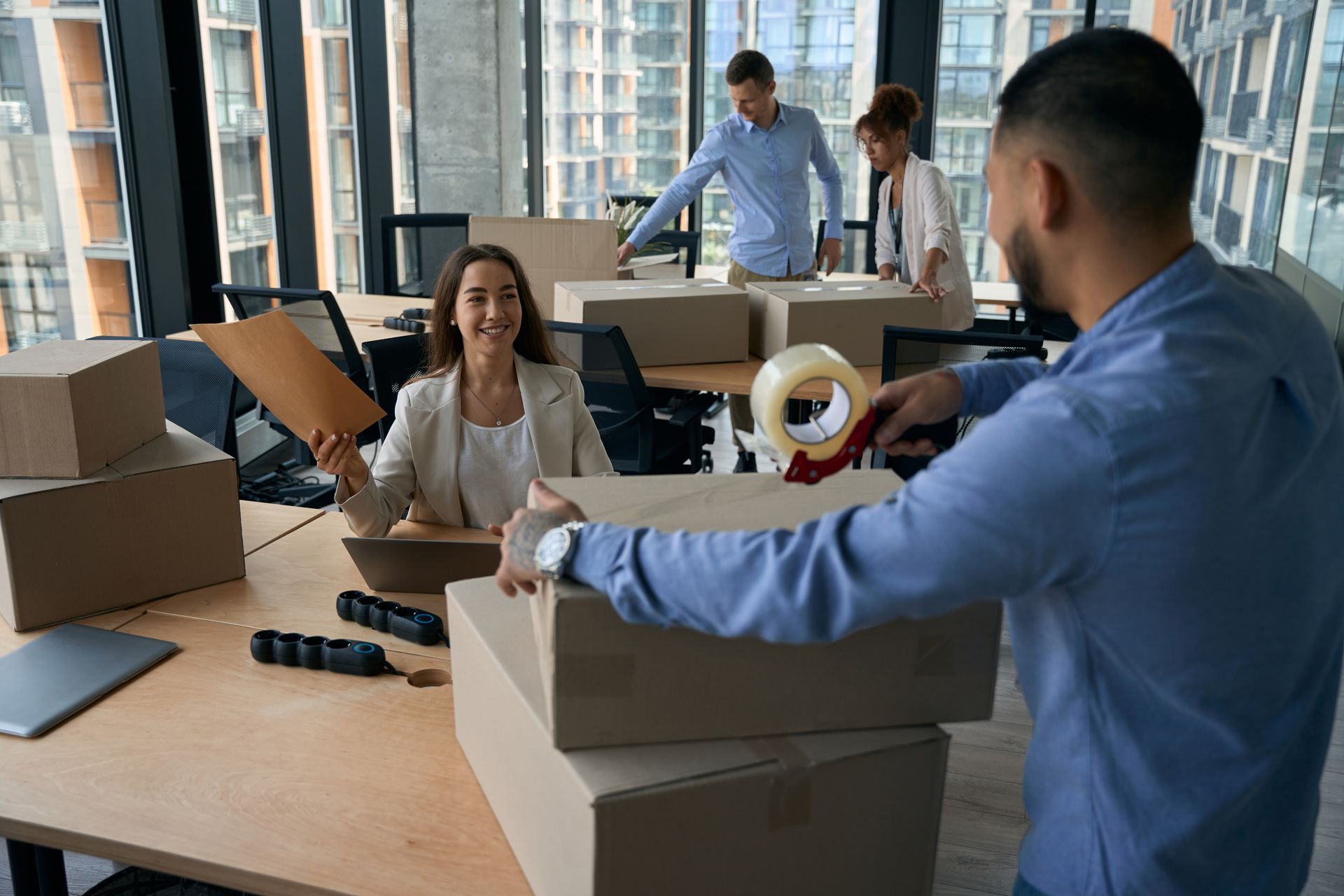 Des employés de bureau emballent des cartons dans un espace de travail lumineux, une femme utilise un ordinateur portable tandis que d'autres scellent les colis.