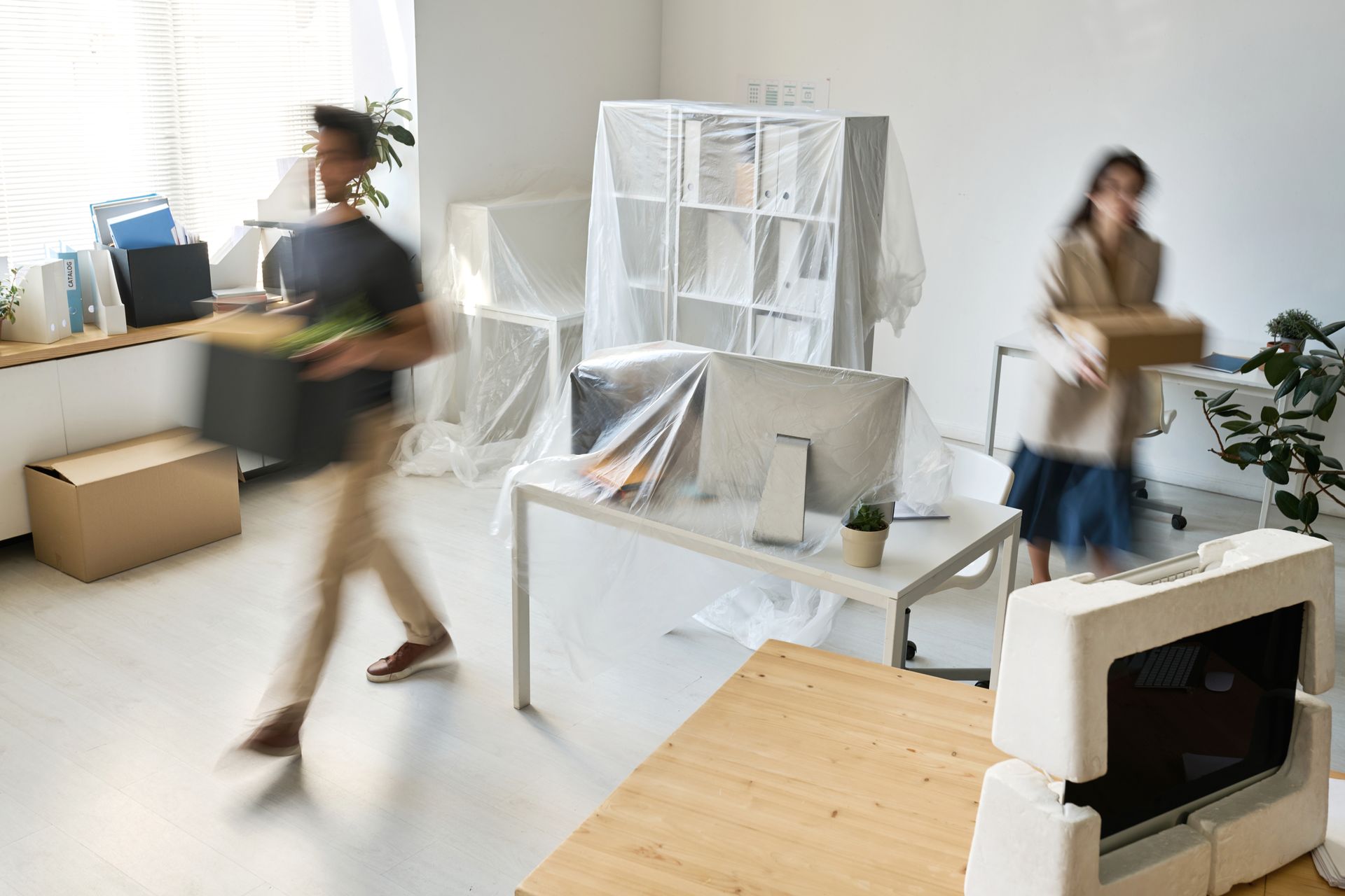 Deux personnes déplacent des cartons dans un bureau lumineux meublé de bureaux et d'étagères lors d'un déménagement.