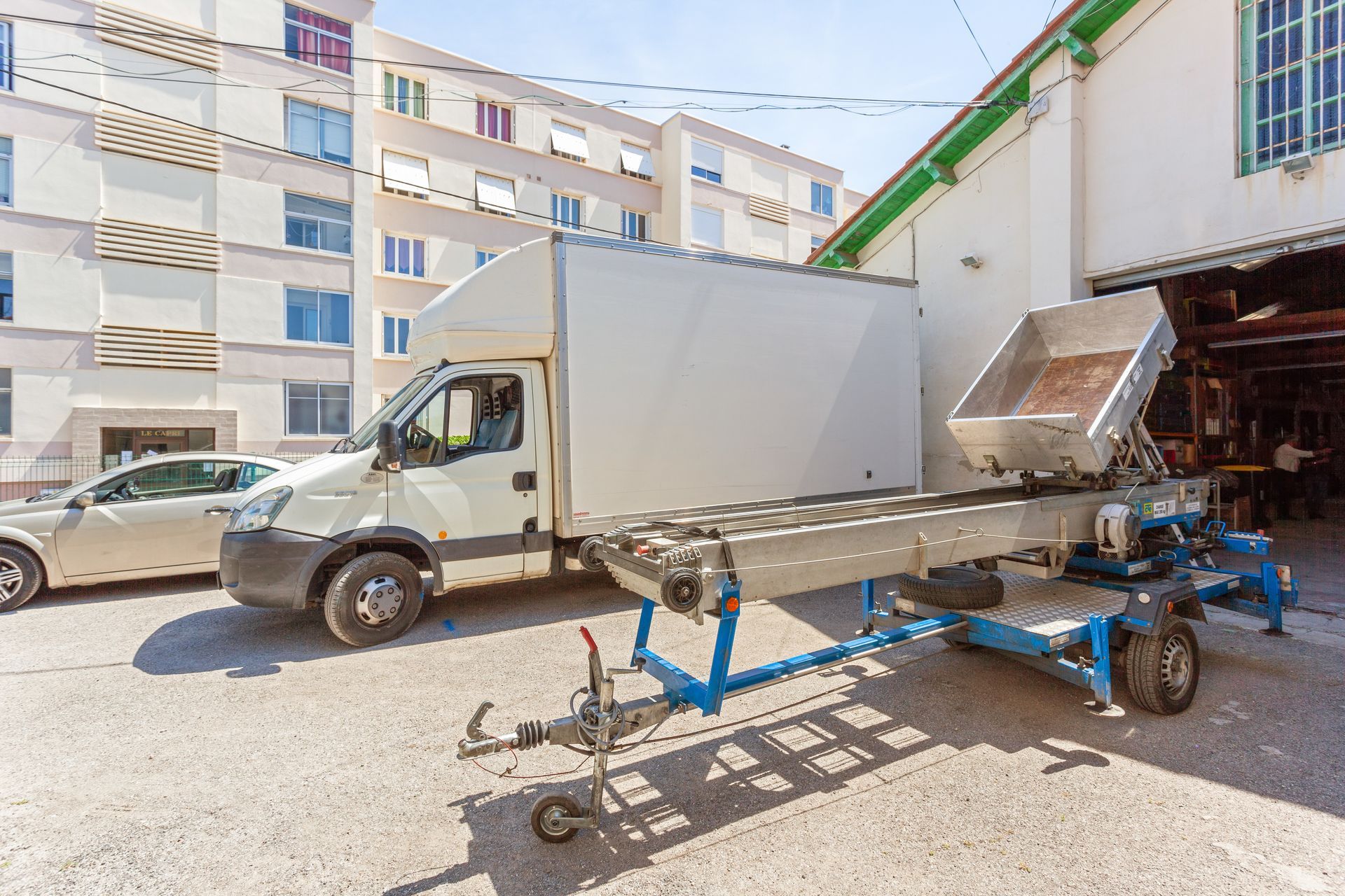 Un camion blanc est stationné dans une cour à côté d'immeubles d'appartements, avec une remorque plateau bleue devant.