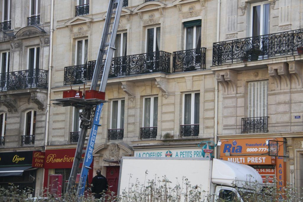 Un ouvrier se trouve sur une nacelle élévatrice rouge à côté d'un camion blanc devant un immeuble d'appartements avec balcons.
