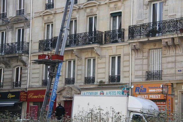 Vue au niveau de la rue de la façade d'un immeuble parisien avec balcons, enseignes de magasins et camion utilitaire en contrebas.