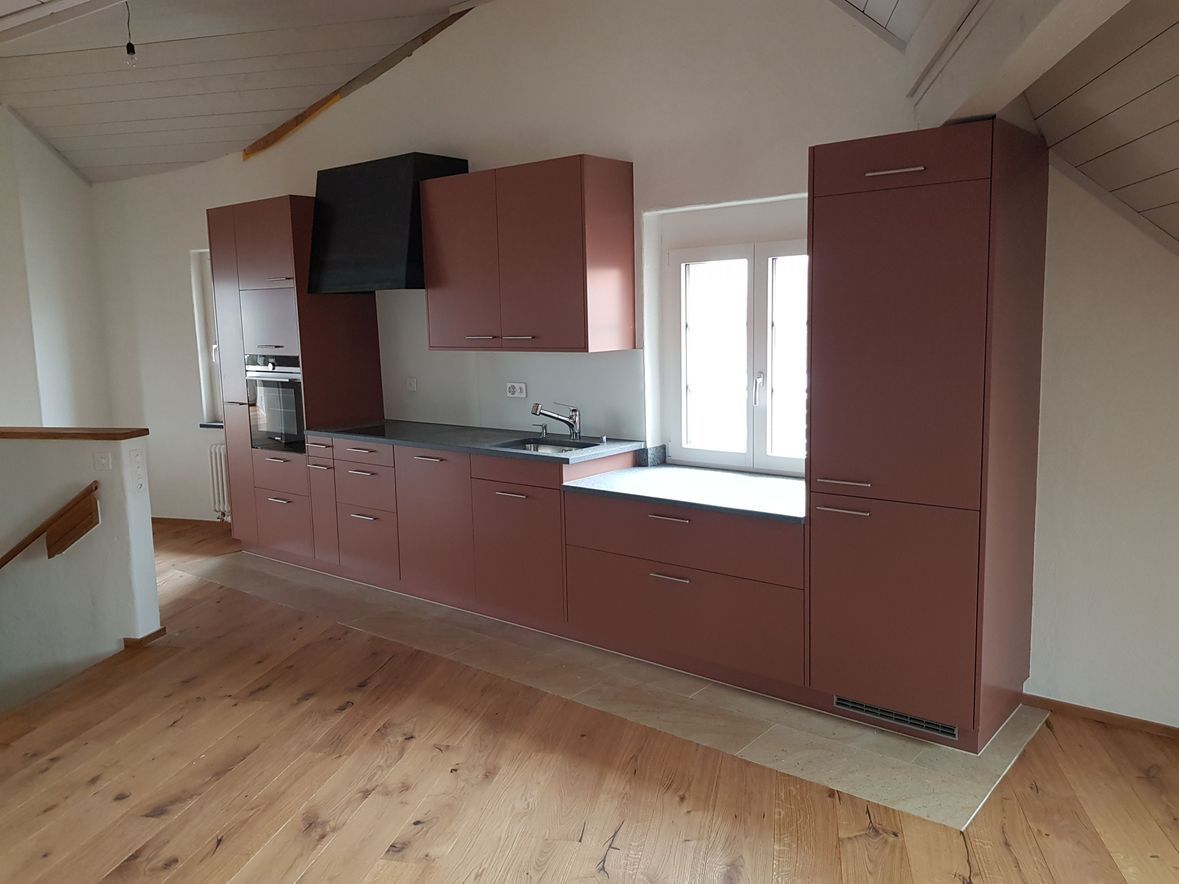 Küche mit bordeauxroten Schränken, Edelstahlgeräten und einem Fenster. Holzböden und Dachschräge.