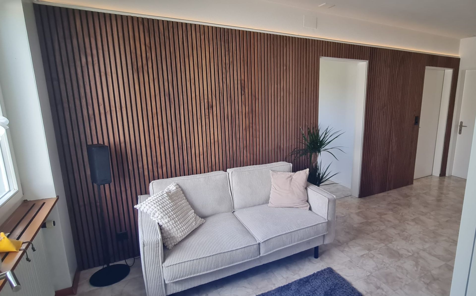 Ein kleines Wohnzimmer mit grauem Sofa, hölzerner Akzentwand und zwei weissen Türen.