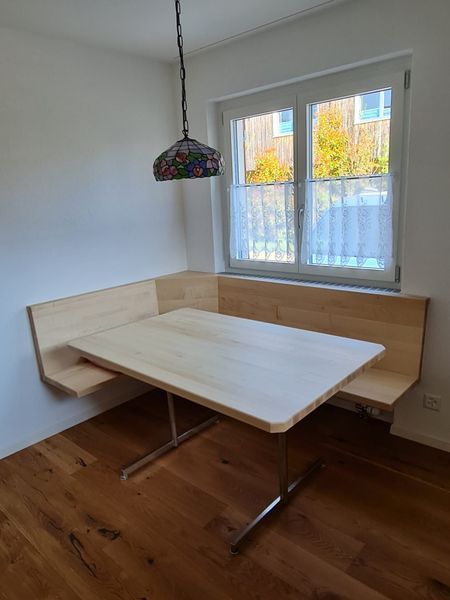 Eingebaute Eckbank aus Holz mit Tisch vor dem Fenster. Darüber eine Pendelleuchte.